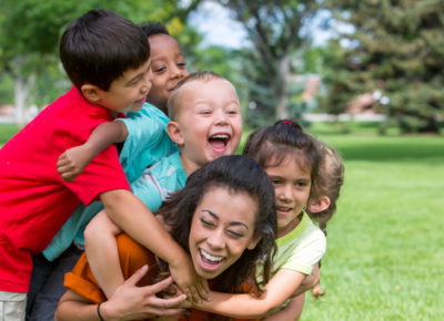 A group of four children laughing as they climb on their children's ministry volunteer.