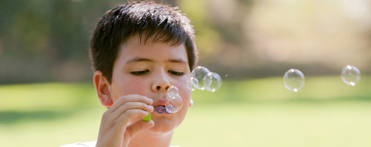 An elementary boy is outside blowing bubbles.