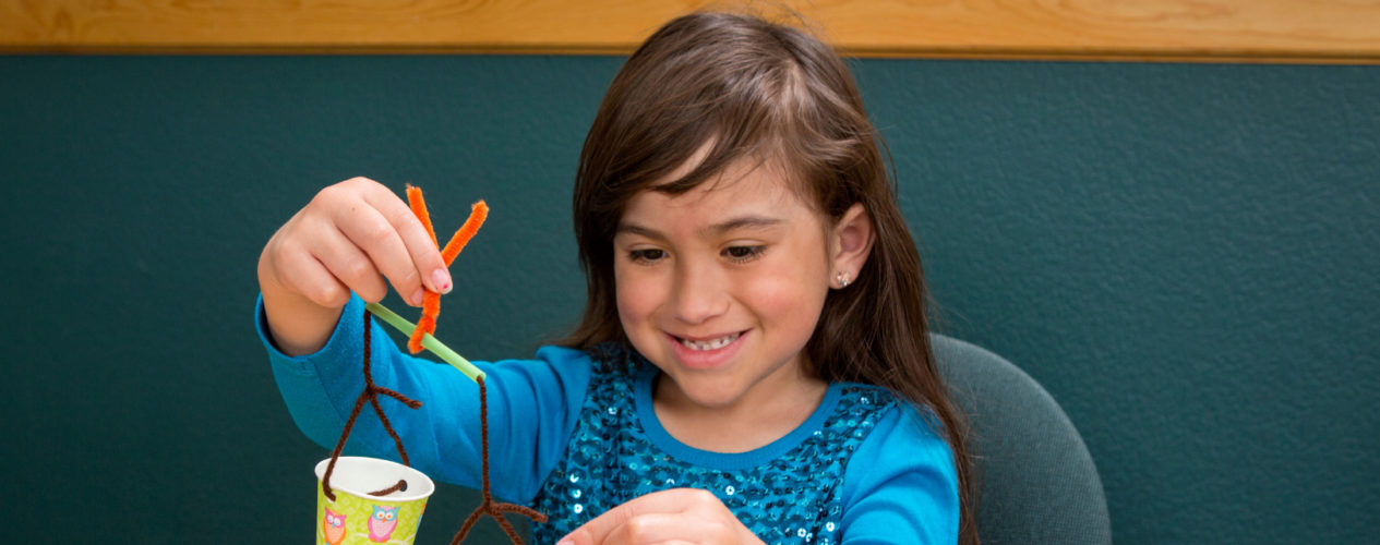 An elementary girl is sitting at a table making a wind chimes.