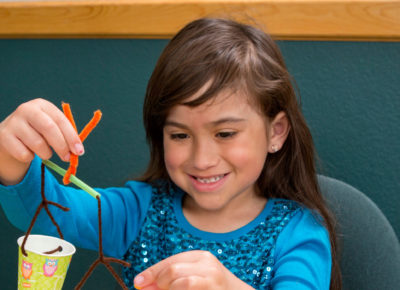 An elementary girl is sitting at a table making a wind chimes.