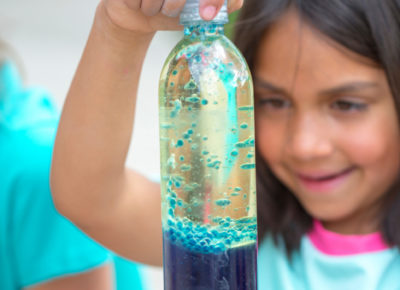 An elementary girl hols a bottle with oil and water in it. The water is blue.