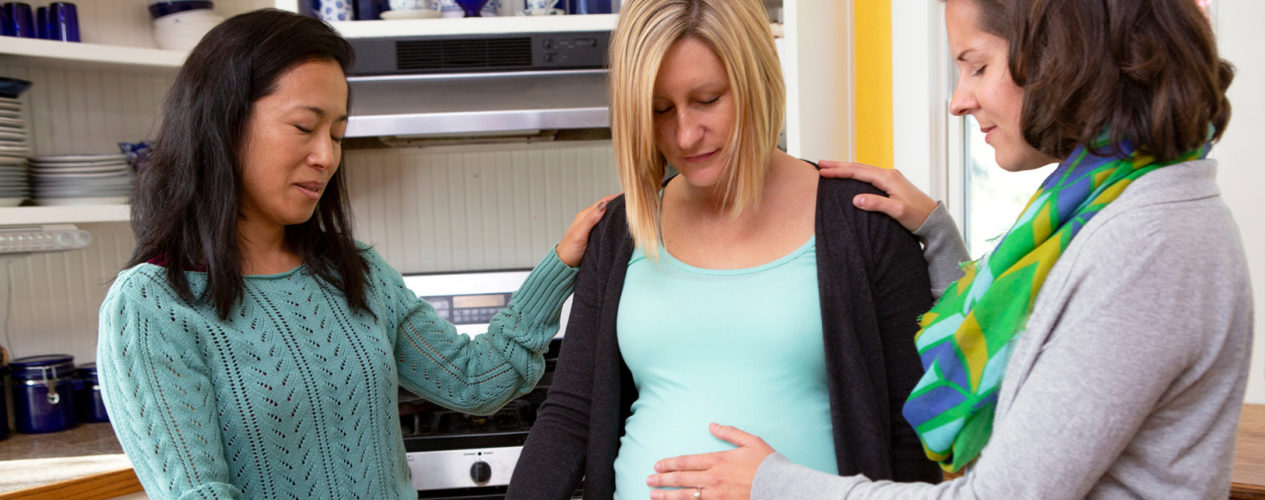 Two woman pray over a very pregnant woman. They are standing in a kitchen.