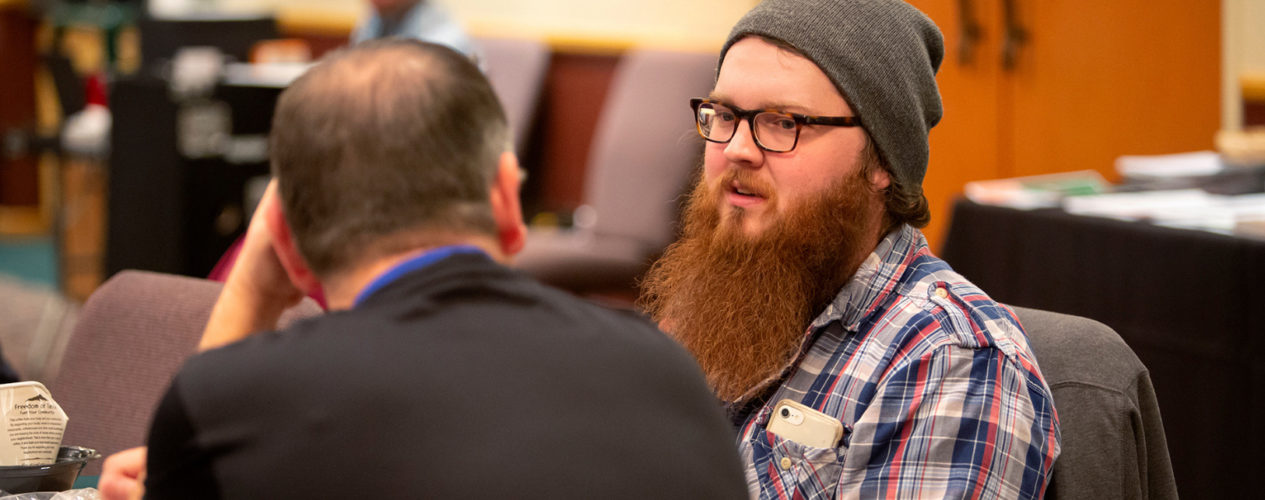 A man with a long beard and beanie hat talks with another man sitting at a table.