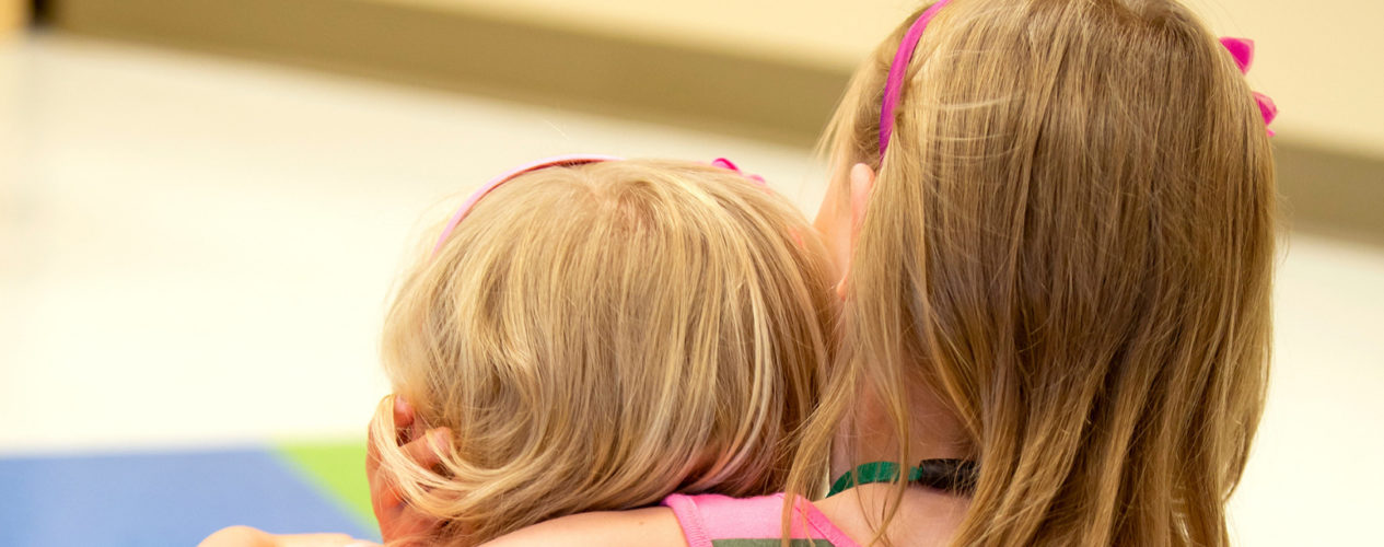 Two girls with blonde hair are sitting on the floor. The older one, who's about 7, is hugging the younger one, who is a preschooler.