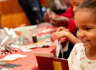 Preschool girl smiles as she sits at a table of crafts.