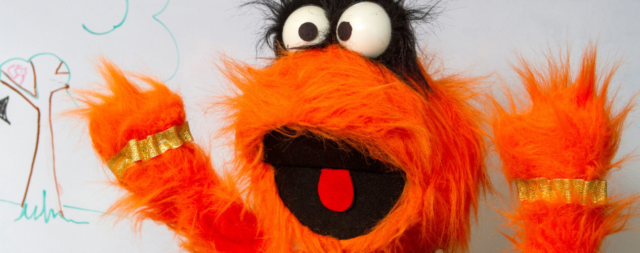 A bright orange monster puppet is standing in front of a white board.