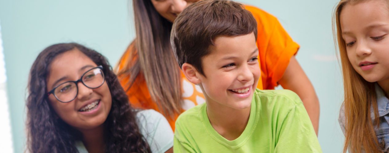 A preteen boy smiles as he looks towards his left. There are two preteen girls sitting with him and one female volunteer.