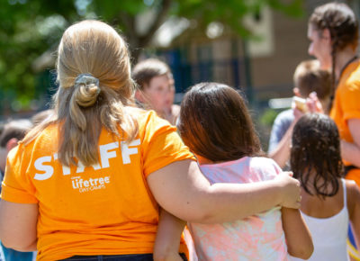 A camp counselor in a bright orange shirt that says "staff" on the back has her arm around a preteen girl.