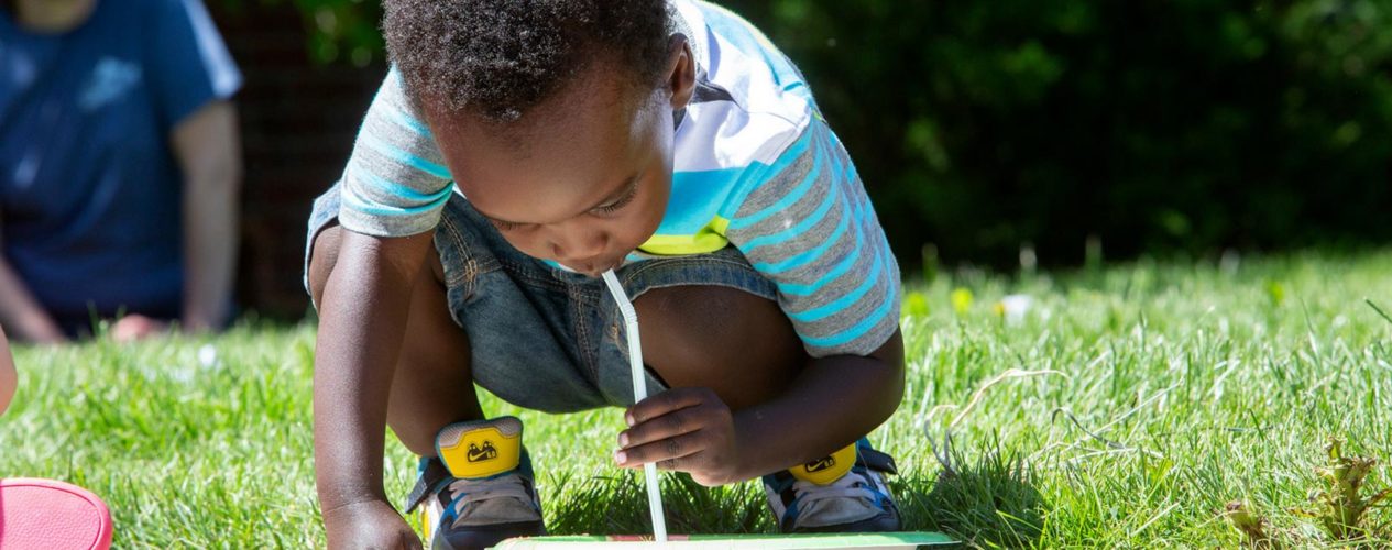 Little boy, squatting in front of a paper play, is using a drinking straw to move something off the plate.