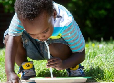 Little boy, squatting in front of a paper play, is using a drinking straw to move something off the plate.