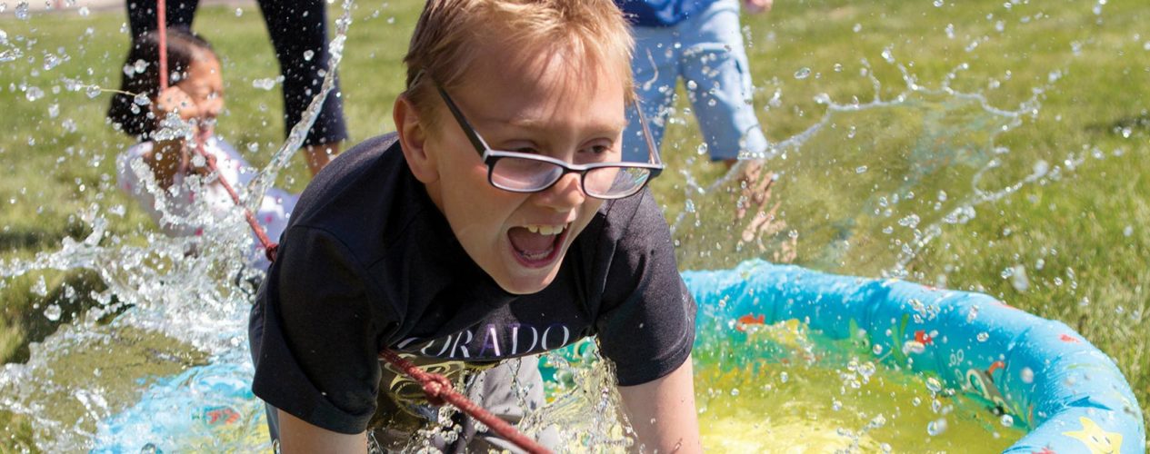 A boy is falling into a kiddie pool, splashing water everywhere. He's got a huge smiling and his glasses are sliding off his face.