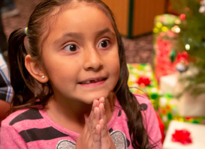 Elementary aged girl folds her hands in prayer excitedly. She's near a Christmas tree filled with presents.