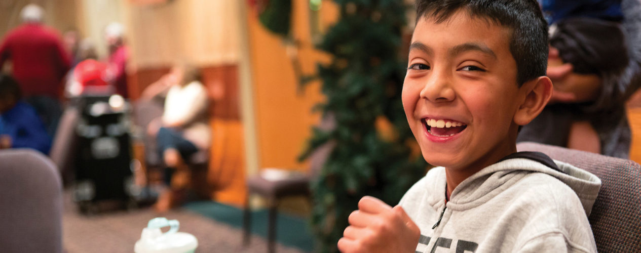 Preteen boy smiles as he participates in a craft. The room he is in is festively decorated for Christmas.