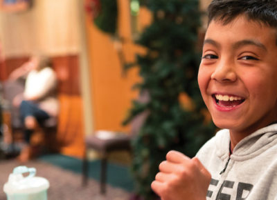Preteen boy smiles as he participates in a craft. The room he is in is festively decorated for Christmas.