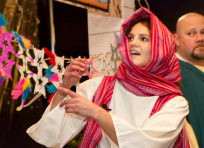 Young female volunteer in a white robe and ink and red head scarf. She's pointing off screen with her mouth open. There's a middle-aged bald man wearing Biblical clothing as well. They are in front of a marketplace with cardboard sunglasses.