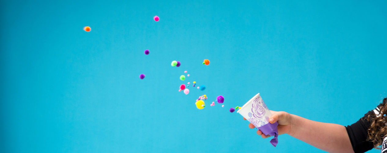 A paper cup with a ballon on the bottom launches colorful pom poms out in front of a bright blue background.