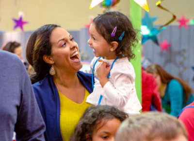 A mom and preschool daughter joyfully smile. They are surround by other families at an Easter event.