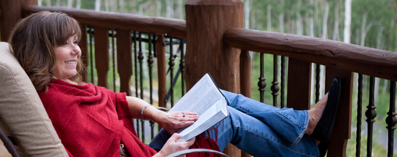 Woman reading her Bible on a patio. She's in a cozy chair with her feet propped up.