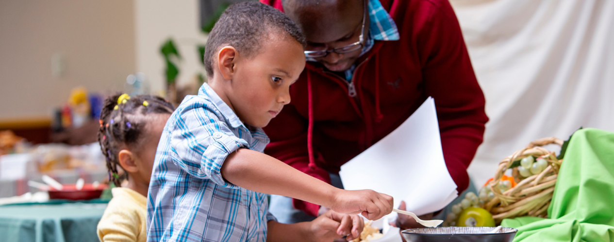 Dad helps elementary-aged boy scoop food out of a bowl with a spoon.