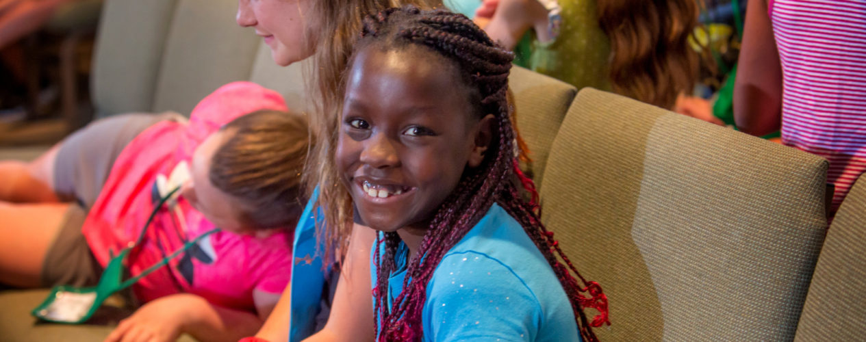 A preteen girl is sitting in a sanctuary chair. She is smiling straight at the camera.
