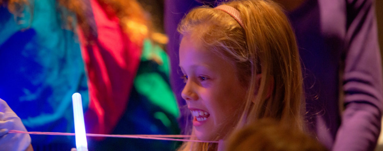 An elementary-aged girl is grinning widely as see looks at a glow stick in her hand.