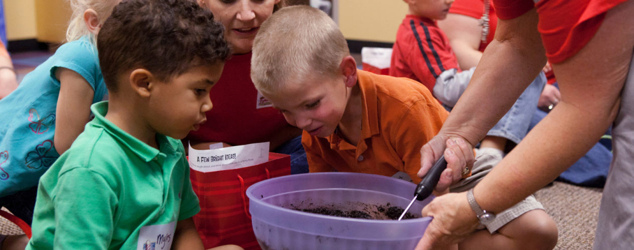 A group of children gather around a mixing bowl that an adult is holding. They are backing a Scripture cake.