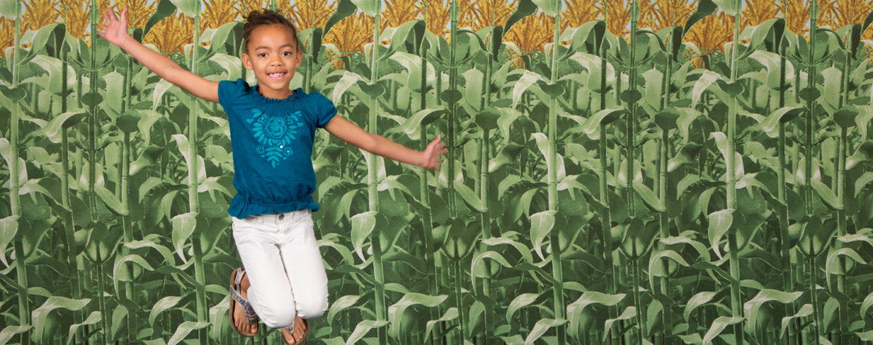 An elementary-aged girl leaps up for a photo in front of a corn backdrop.