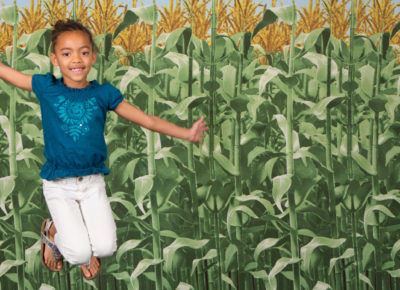 An elementary-aged girl leaps up for a photo in front of a corn backdrop.
