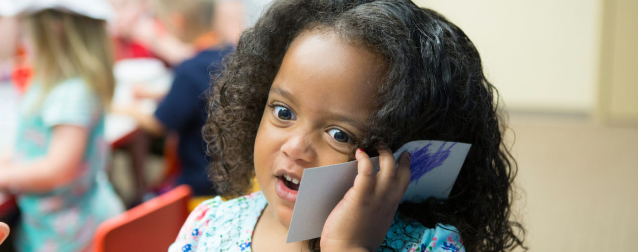 A preschool girl holds up a rectangle piece of card stock to her ear like she's on the phone.