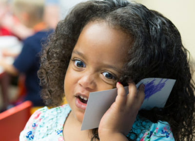 A preschool girl holds up a rectangle piece of card stock to her ear like she's on the phone.
