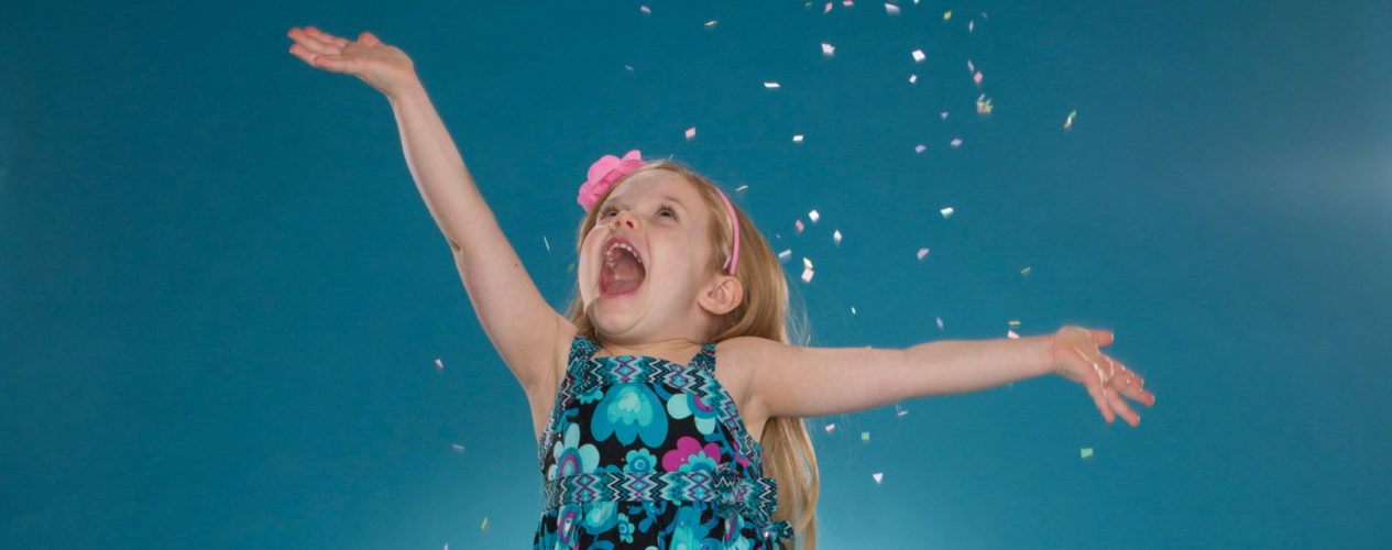 Kindergarten girl wearing a blue dress and pink flowered headband is jumping for joy as confetti rains down.