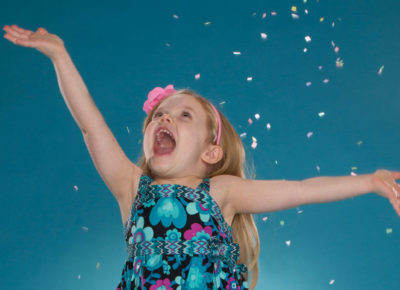 Kindergarten girl wearing a blue dress and pink flowered headband is jumping for joy as confetti rains down.