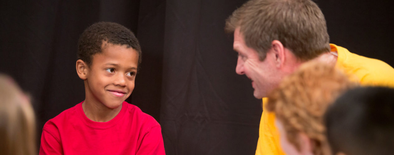 Male volunteer in a bright yellow shirt talks to a third grade boy in a bright red shirt. The boys has a smirk on his face.