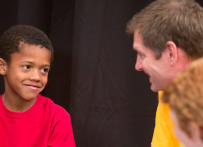 Male volunteer in a bright yellow shirt talks to a third grade boy in a bright red shirt. The boys has a smirk on his face.