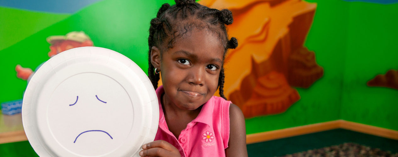 A preschool girl holds up a paper plate with a sad face on it. She's making the same face.