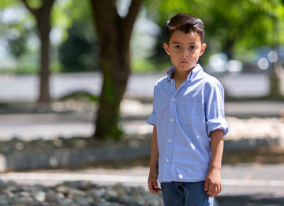 An elementary-aged boy standing outside alone. He's looking directly into the camera with a serious expression.