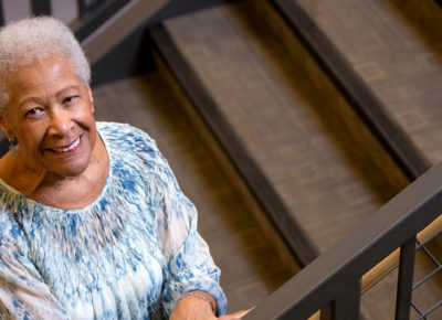 Elderly woman with a welcoming smile stands on a flight of stairs.