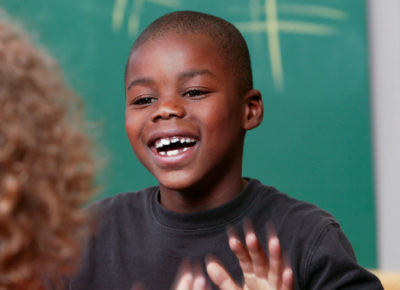 Elementary boy sitting in front of a chalk board, playing a hand clapping game with a elementary girl who's back is towards the camera. She has very curly hair.