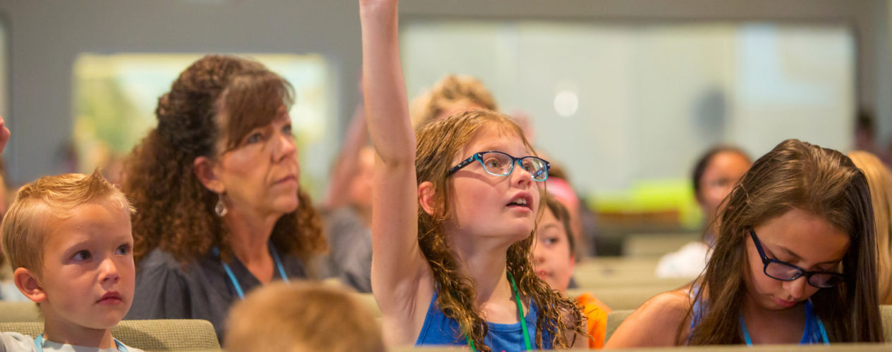 An elementary-aged girl raising her hand in a church sanctuary.