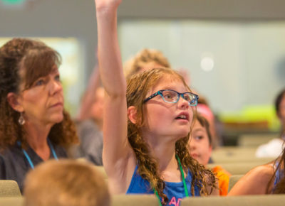 An elementary-aged girl raising her hand in a church sanctuary.