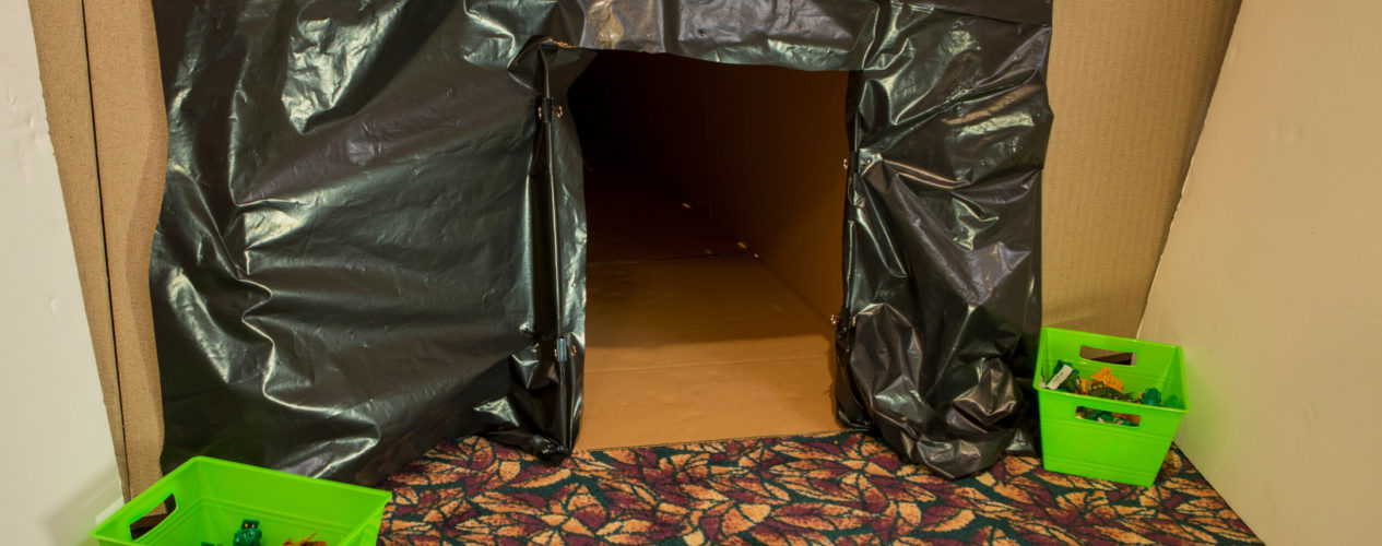 The entrance of a tunnel made of cardboard boxes is surrounded by black trash bags.