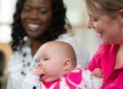 A few women are gathered. One woman has her baby sitting on her lap as other women smile at the baby.
