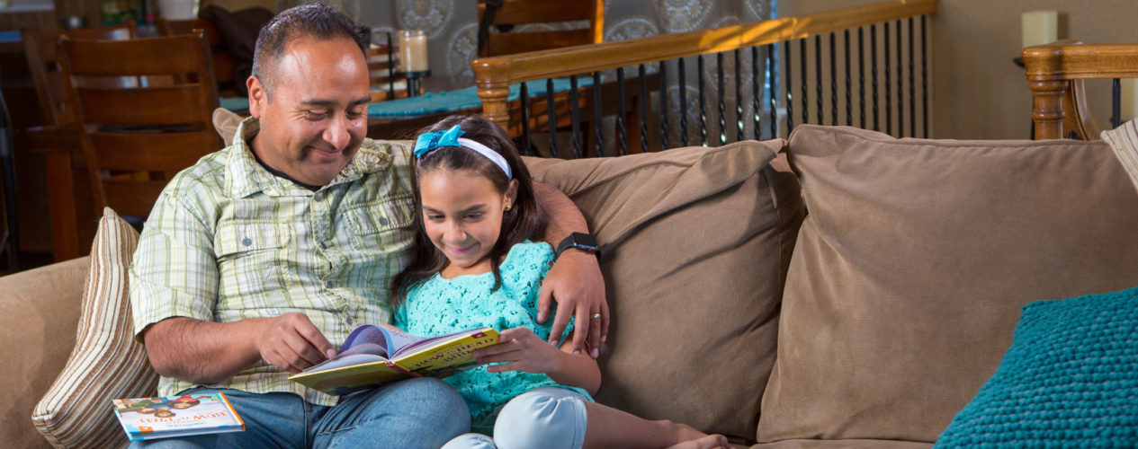 A dad and daughter sit on their couch reading books.