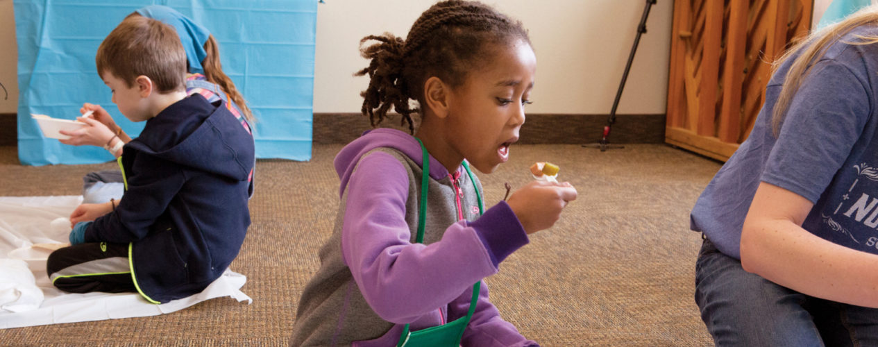 An elementary girl sitting with other kids in a circle on the ground. She's eating a snack with a spoon.
