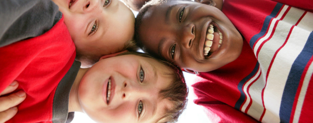 A group of elementary-aged boys huddled shoulder-to-shoulder, looking down at a camera that sits between them. They have huge smiles on their faces.