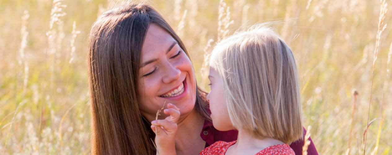 A mom smiles at her young daughter as the daughter sits on her lap in a field.