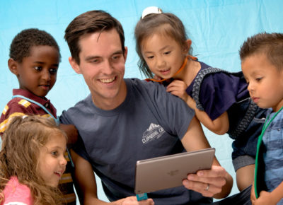 A male volunteer has four kids around him looking at the iPad he's holding.
