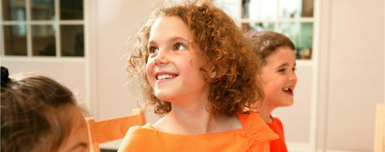 A young girl is sitting at a table with two friends. She's looking up and over her shoulder and smiling at her teacher.