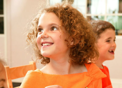A young girl is sitting at a table with two friends. She's looking up and over her shoulder and smiling at her teacher.