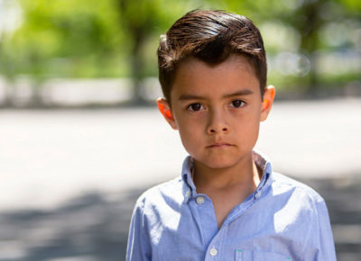 Elementary age kid standing in a parking lot. He's wearing a button up shirt and is looking directly at the camera with a very serious face.